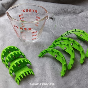 May include: A clear Pyrex measuring cup with red markings and a handle sits next to several bright green kitchen gadgets. The gadgets have a curved design with small, pointed protrusions.