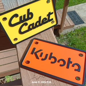 May include: Two rectangular signs with black lettering. One is yellow with "Cub Cadet" and the other is orange with "Kubota". Both have black borders and mounting holes. The date "June 21, 2025 17:15" is visible.