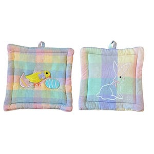 Puede incluir: Dos agarraderas cuadradas de colores pastel. Una presenta un pollito amarillo bordado saliendo de un huevo y un huevo azul. La otra tiene un conejito blanco bordado. Ambas tienen un lazo para colgar.