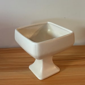 May include: A cream-coloured ceramic pedestal bowl with a square-shaped basin and a matching square base. The vessel features smooth, clean lines and a glossy finish, standing upright with a flared stem connecting the bowl to the base.