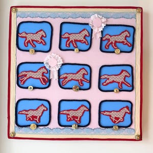 以下が含まれることがあります： 青い背景に赤と白で刺繍された9つの馬のシルエットが特徴の、額装されたテキスタイルアート作品。 ピンクと赤の縁取りがあり、金色のボタンとレースのディテールで飾られています。 装飾的なロゼットも含まれています。