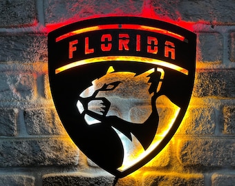 May include: Black metal wall art featuring the word "FLORIDA" and a panther logo, illuminated with red and yellow LED lights. The shield-shaped sign is mounted on a brick wall, creating a striking visual effect.