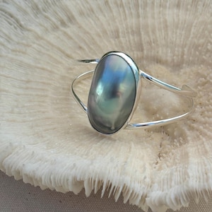 May include: A silver cuff bracelet featuring a large, oval-shaped, iridescent gray pearl set in a smooth metal bezel. The band is constructed from two thin, polished silver wires that meet at the sides to hold the central gemstone securely.
