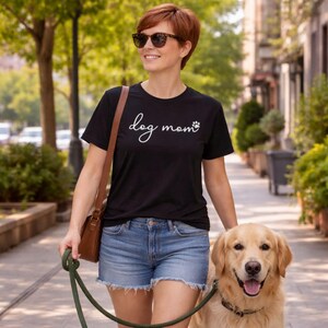 Könnte beinhalten: Schwarzes T-Shirt mit dem Schriftzug "dog mom" in Schreibschrift und einem Pfotenabdruck. Das Shirt wird mit Jeansshorts kombiniert. Ein Golden Retriever ist an der Leine. Die Person trägt eine Sonnenbrille und eine braune Tasche.