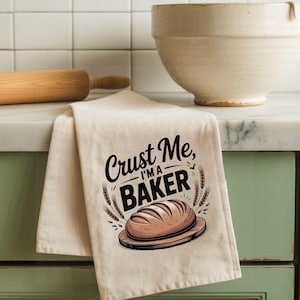 May include: A beige kitchen towel with the text "Crust Me, I'm a Baker" in black script. The design includes an illustration of a loaf of bread on a wooden board, surrounded by wheat stalks. A wooden rolling pin and a large white bowl are in the background.