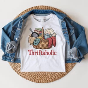 Pode incluir: Camiseta branca com um gráfico de uma cesta de vime cheia de livros, uma chaleira, um prato e uma frigideira de ferro fundido. A cesta contém um cachecol de malha azul e vermelho. A palavra "Thriftaholic" está impressa em vermelho abaixo.