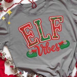 May include: A gray t-shirt with the words "ELF Vibes" in red and green. The letters "ELF" are in a plaid pattern with a white outline. The word "Vibes" is in a cursive font, with elf shoe accents.