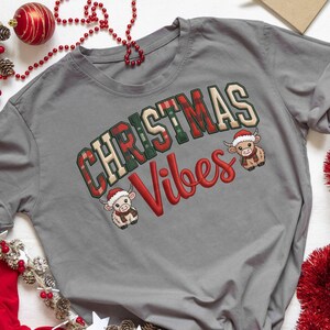 Könnte beinhalten: Graues T-Shirt mit dem Schriftzug "CHRISTMAS Vibes" in rot-grün karierten Buchstaben. Zwei Cartoon-Kühe mit Weihnachtsmützen sind auf dem Shirt abgebildet. Festliche Weihnachtsbekleidung.