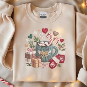 Op de afbeelding: Beige sweatshirt met een geborduurd winterontwerp. Het ontwerp omvat een blauwgroene mok warme chocolademelk met marshmallows, zuurstokken, wanten, ingepakte cadeaus en harten. De sweatshirt heeft een ronde hals en lange mouwen.