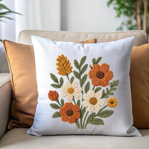 Può includere: Cuscino quadrato bianco con un ricamo floreale. Il design presenta fiori arancioni, gialli e color crema con foglie verdi. Il cuscino è posto su un divano beige con altri due cuscini.