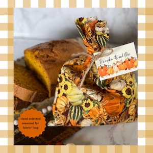 May include: A fabric bag featuring a pattern of orange pumpkins, yellow sunflowers, and green gourds on a dark background. The bag is tied with twine and has a white tag attached that reads Pumpkin Bread Mix with pumpkin illustrations.