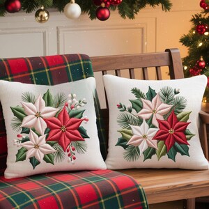 May include: Two white square throw pillows with embroidered poinsettias in red and pink, surrounded by green leaves and pine needles. The pillows rest on a wooden bench with a red and green plaid blanket.