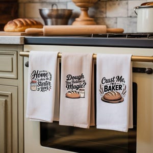 Könnte beinhalten: Drei Küchentücher mit Backmotiven und Illustrationen hängen an einem Herd. Die Handtücher sind weiß und beige. Eines sagt "Home is where the starter lives", ein anderes "Dough, You Believe in Magic?" und das letzte "Crust Me, I'm a Baker."
