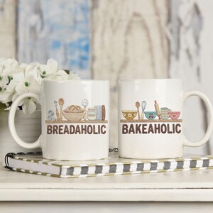 May include: Two white ceramic mugs with handles, each featuring a brown shelf with baking-related illustrations. One mug says "BREADAHOLIC" and the other says "BAKEAHOLIC" in brown text. The mugs are on a striped notebook.