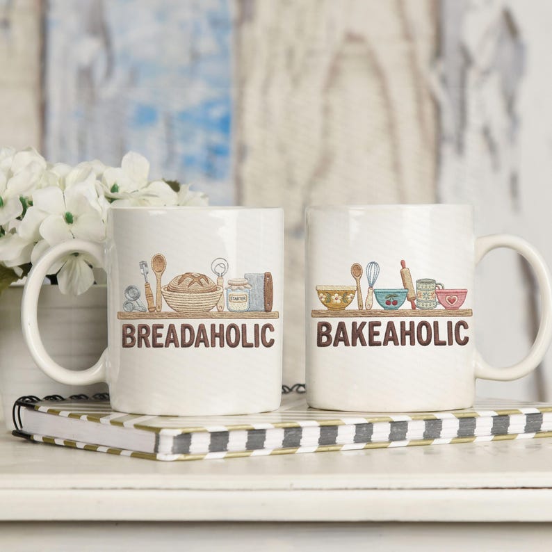May include: Two white ceramic mugs with handles, each featuring a brown shelf with baking-related illustrations. One mug says "BREADAHOLIC" and the other says "BAKEAHOLIC" in brown text. The mugs are on a striped notebook.