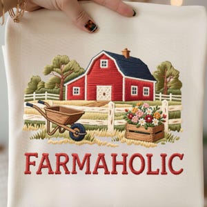 May include: Cream-coloured embroidered farmhouse-themed towel featuring a red barn with a blue roof, a wooden wheelbarrow, and a wooden crate filled with flowers. The text "FARMAHOLIC" is embroidered in red.