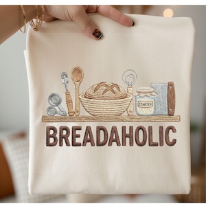 以下が含まれることがあります： パン作りの道具とパンの刺繍デザインが施されたクリーム色のスウェットシャツ。「BREADAHOLIC」の文字が茶色の文字でデザインの下に刺繍されています。