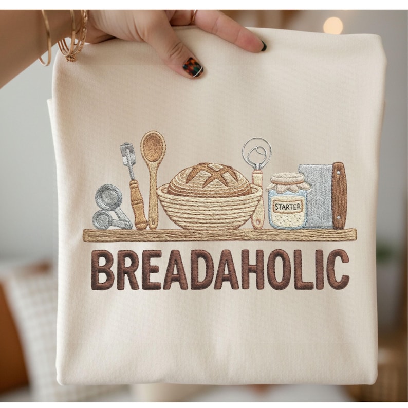 May include: Cream-colored sweatshirt with an embroidered design featuring bread-making tools and a loaf of bread. The word "BREADAHOLIC" is embroidered in brown letters below the design.