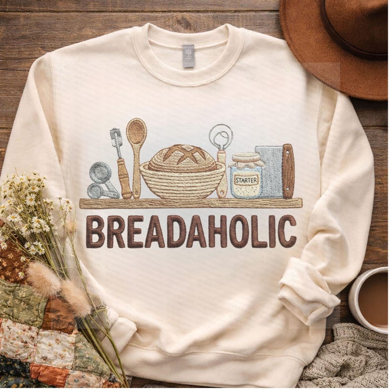 May include: Cream-colored sweatshirt with a graphic of bread-making tools and a loaf of bread on a shelf. The word "BREADAHOLIC" is printed in brown block letters below the shelf.