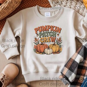 Könnte beinhalten: Cremefarbenes Sweatshirt mit dem Text "PUMPKIN PATCH CREW" in Orange, Grün und Gelb. Die Grafik zeigt drei Kürbisse in Orange, Weiß und Gelb, mit Herbstlaub. Ein gemütliches Herbst-Design.