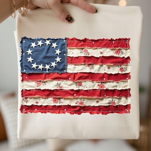 Peut inclure: Un sac fourre-tout crème avec un motif de drapeau américain stylisé. Le drapeau présente un carré bleu avec un cercle d'étoiles blanches et des rayures rouges et crème alternées. Les rayures crème ont un motif floral.