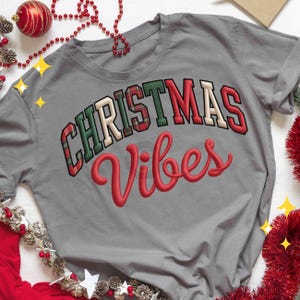 Puede incluir: Camiseta gris con la frase "CHRISTMAS Vibes". La palabra "CHRISTMAS" está en un patrón de cuadros rojos, verdes y blancos. La palabra "Vibes" está en una fuente roja, ideal para la temporada navideña.