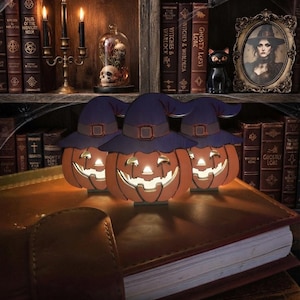 Può includere: Tre decorazioni a forma di zucca arancione con volti intagliati da jack-o'-lantern, ognuna con un cappello da strega viola scuro con fibbia. Le zucche sono illuminate dall'interno, emettono una luce calda e sono disposte su un libro spesso.