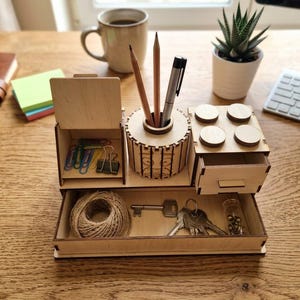 May include: A light-colored wooden desk organizer featuring a central cylindrical pen holder, a small lidded compartment, a drawer with four circular top accents, and a wide bottom tray containing a ball of twine, keys, and small metal pins.