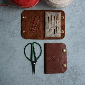 May include: A brown leather sewing kit with a flap that opens to reveal a felt interior with needles and thread. The kit also includes a pair of small green scissors and a separate brown leather pouch with the text "Le Rouge" and "Made by Isabelle B."
