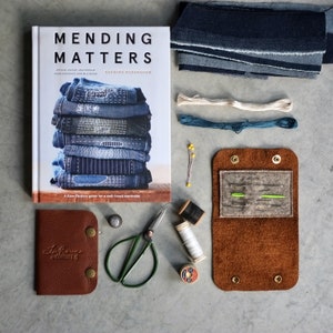 May include: A brown leather sewing kit with a flap that opens to reveal a felt interior with needles and thread. The kit is on a gray surface with a book titled "Mending Matters" and other sewing supplies.