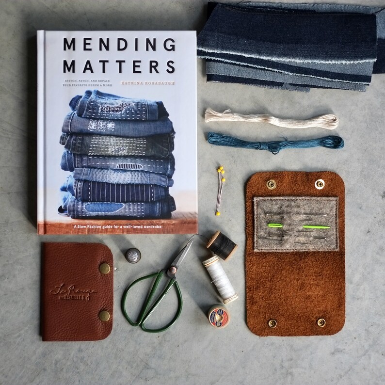 May include: A brown leather sewing kit with a flap that opens to reveal a felt interior with needles and thread. The kit is on a gray surface with a book titled "Mending Matters" and other sewing supplies.