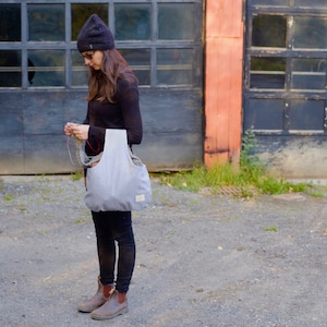 May include: A light gray tote bag with vertical stripes, carried over the shoulder. The bag has a rectangular shape with a wide opening and a small label on the front. The person is wearing a black beanie, sunglasses, and black clothing.