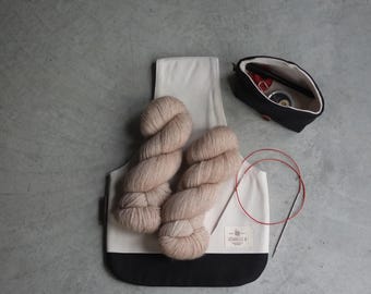 Puede incluir: Un kit de manualidades con dos madejas de hilo marrón claro, un bolso tote blanco y negro, una bolsa negra y agujas de tejer circulares rojas. El bolso tote tiene una etiqueta que dice "Isabelle B."
