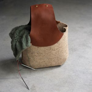 Könnte beinhalten: Ein beigefarbener Filzkorb zur Aufbewahrung von Garn mit einem braunen Ledergriff und einem grünen Strickpullover im Inneren. Zwei Stricknadeln liegen auf dem Boden vor dem Korb.