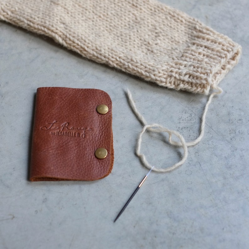 May include: A brown leather needle case with gold metal snaps and the text "Le Rouge" and "Cire & Sabellerie" embossed on the front. A white yarn and a metal needle are on a gray surface.
