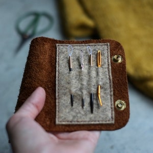 May include: A brown leather case with a flap closure, containing six needles with different colored threads. The case has two metal snaps on the side.