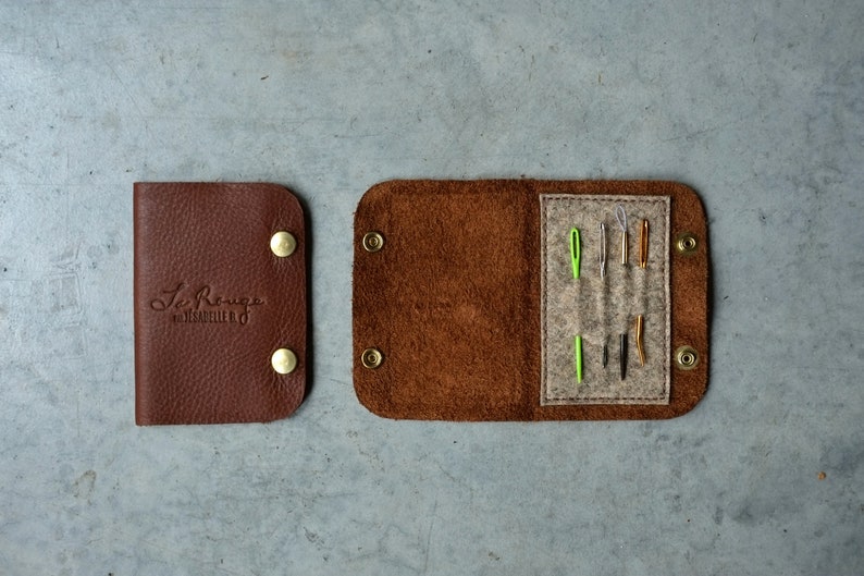 May include: A brown leather needle case with a flap that opens to reveal a felt interior with several needles. The case has a brass snap closure and the text "Le Rouge" is embossed on the front.