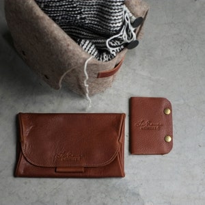 May include: Two brown leather wallets with the text "Le Rouge Habilleur" embossed on them. The wallets are on a gray surface.