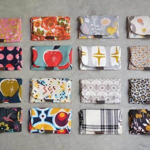 May include: A collection of sixteen fabric wallets with various patterns, including floral, fruit, geometric, and animal prints. Each wallet has a flap closure and a small brown leather accent. The wallets are arranged on a gray surface.