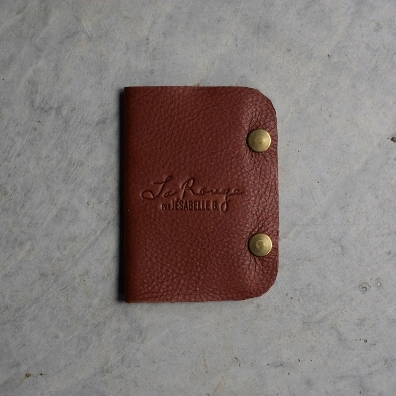 May include: A small, brown leather wallet with the text "La Rouge by Isabelle B." embossed on the front. The wallet has two brass snaps on the right side.