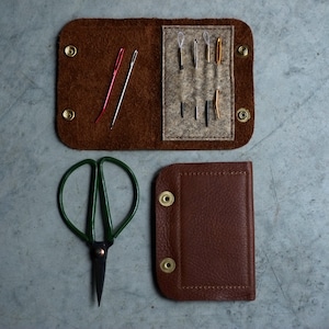 May include: A brown leather sewing kit with a variety of needles and a pair of green scissors. The kit has a flap that closes with a snap.