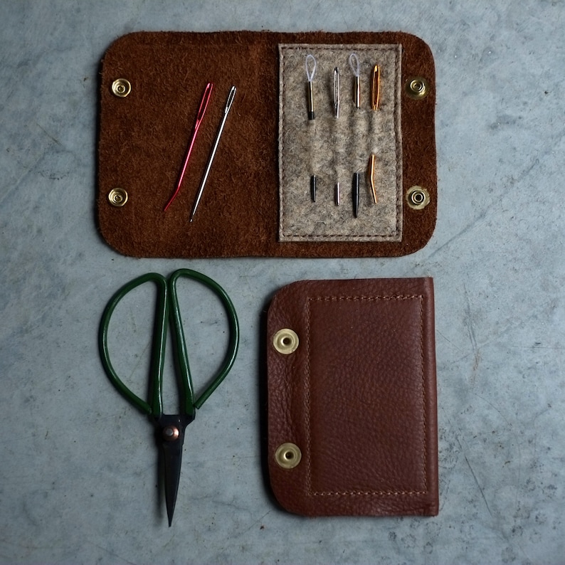 May include: A brown leather sewing kit with a variety of needles and a pair of green scissors. The kit has a flap that closes with a snap.