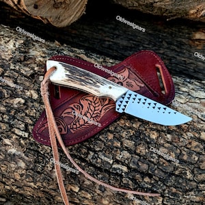 May include: A fixed-blade knife with a textured antler handle and a silver-coloured steel blade featuring a repeating pattern of small black triangles. The knife includes a brown leather sheath with embossed decorative details and a leather lanyard.