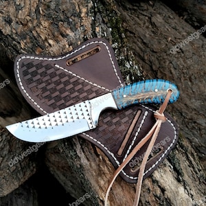 May include: A handcrafted knife with a patterned blade and a blue and brown handle, resting in a brown leather sheath. The sheath features a woven design and white stitching, with leather ties. The knife and sheath are displayed on a textured wooden surface.