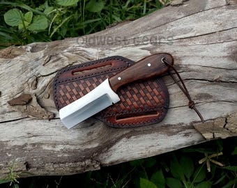 Peut inclure: Un couteau artisanal avec un manche en bois foncé et une lame argentée repose sur un étui en cuir marron. L'étui présente un motif tissé et un cordon en cuir. Le couteau et l'étui sont présentés sur une surface en bois vieilli.