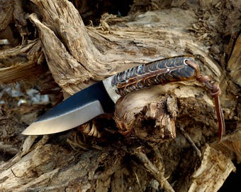 Peut inclure: Un couteau artisanal avec une lame sombre et un manche présentant un motif unique marron et blanc. Le manche est en bois et en os, avec une lanière en cuir attachée. Le couteau repose sur un morceau de bois vieilli.