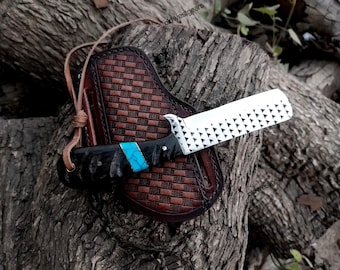 Puede incluir: Un cuchillo artesanal con una hoja blanca con un patrón geométrico y un mango negro con una incrustación turquesa. El cuchillo descansa sobre una funda de cuero marrón con un diseño tejido y una correa de cuero.