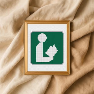 Op de afbeelding: Een ingelijste kruissteek van een bibliotheeksymbool. De afbeelding toont een wit figuur dat een boek leest op een groene achtergrond. De lijst is goudkleurig en de achtergrond is een beige stof.