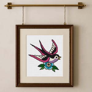 May include: Framed cross-stitch art featuring a traditional swallow tattoo design. The bird is depicted in black, pink, and white, with a blue flower below. The artwork is displayed in a dark brown wooden frame, hanging from a brass rod.