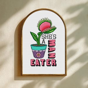 May include: A framed art print featuring a Venus flytrap illustration. The plant is in a teal pot with a purple flower design. The text reads "She's A Man Eater" in pink and black block letters.
